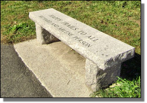 A photo of a granite bench along the Milford Upper Charles trail. The engraving on the bench reads 'Happy Trails to all Richard and Arlene Person'