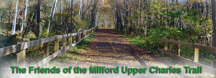 A view down a section of a paved trail passing through a wooded area in the fall. The words 'The Friends of the Milford Upper Charles Trail' are overlayed in green text.